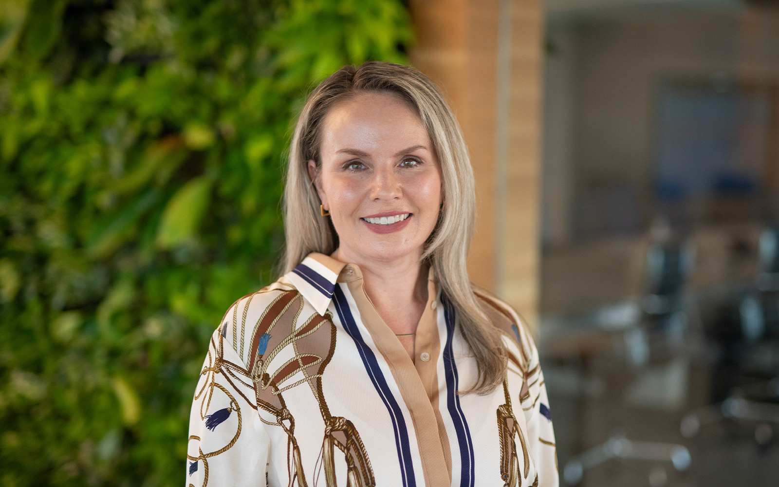 Portrait of a smiling woman with blonde hair wearing a patterned blouse, standing in front of a green wall indoors
