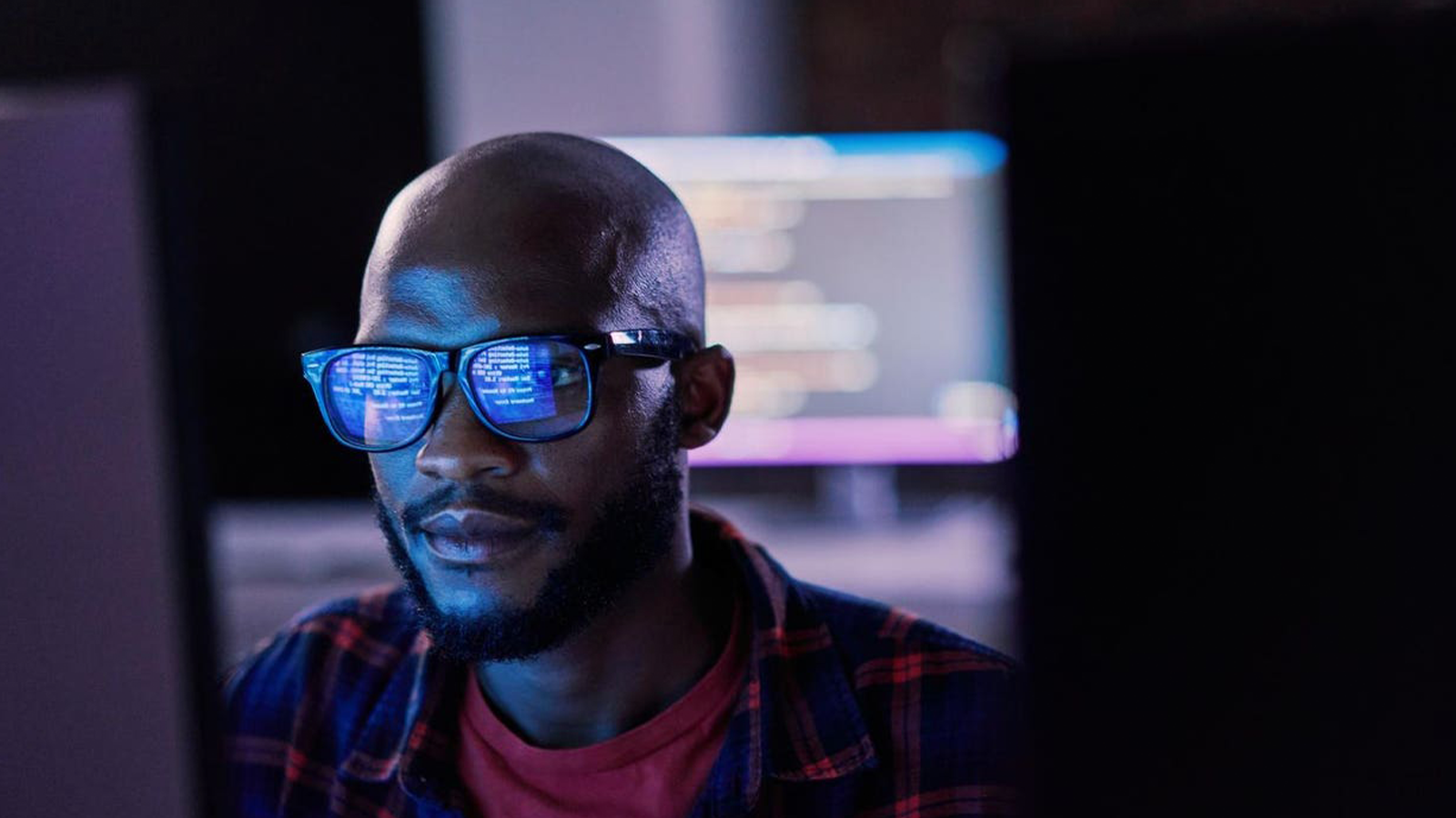 Man with a beard and blue-tinted glasses coding at a dark desk with a glowing monitor behind him.
